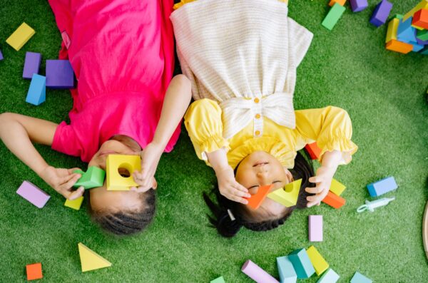 Two children laying on a green turf each holding colorful wooden blocks over their faces.